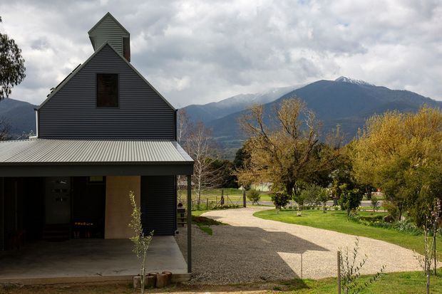 Exterior view of a dark horizontal-clad house with a steep gabled roof, prominent upper gable/tower, and a broad covered carport or entry canopy. A gravel driveway and landscaped garden lead toward the home, with dramatic mountain scenery and autumn trees in the background.