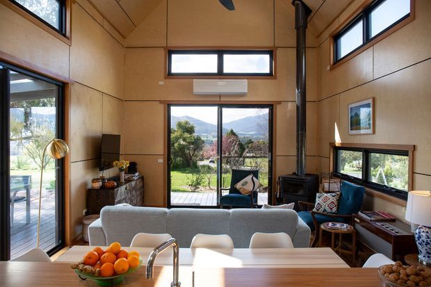 A warm, compact open-plan living area viewed from a kitchen island, featuring timber-lined walls and ceiling, large windows and glazed doors, a grey sofa, a compact dining area, a freestanding black wood-burning stove, and views to a deck, garden, and mountainous landscape. The room has abundant natural light and a strong connection to the outdoors.