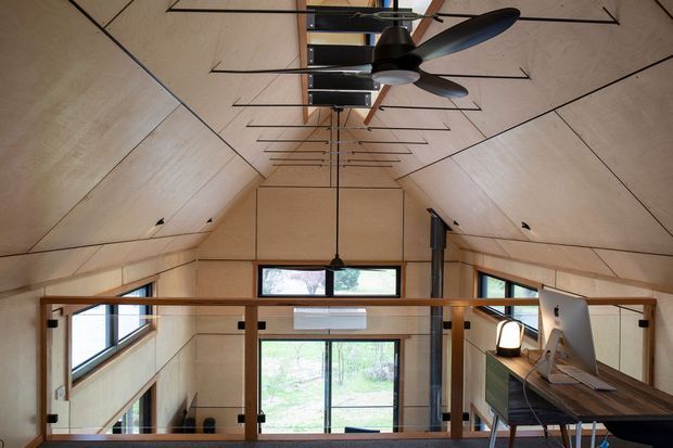 Interior view of a compact contemporary timber-lined space with a steep pitched ceiling, exposed plywood wall and ceiling panels, and a central ceiling fan. A mezzanine or loft level is enclosed by a timber-framed glass balustrade, with large windows and a glazed door bringing in natural light. A built-in or freestanding workspace is visible at right, and a dark vertical flue suggests a heating appliance. The space has a warm, minimalist cabin-like character.