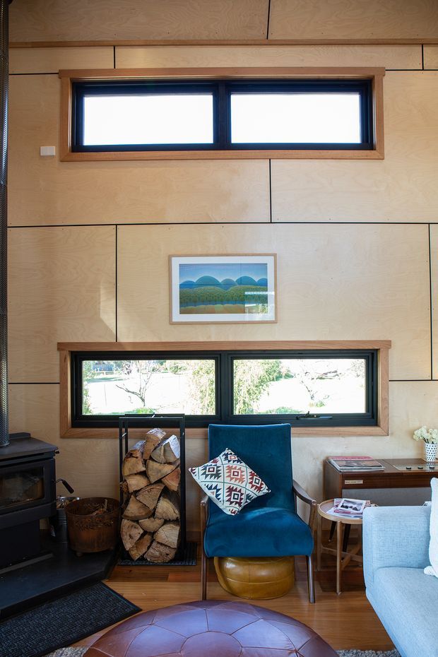 A warm, compact living room lined with plywood wall panels and horizontal black-framed windows. The room features a black wood-burning stove, stacked firewood, a deep teal upholstered armchair with a patterned cushion, a pale sofa, a leather ottoman, and timber flooring. The interior has a cosy cabin-like character with natural materials and strong geometric joinery lines.
