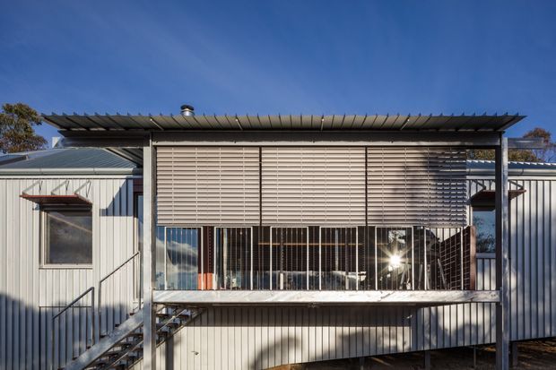 Exterior view of a contemporary elevated building with corrugated metal cladding, a shallow projecting roof, external stairs, and a screened upper-level terrace or balcony. Horizontal louvres shade the glazing, while vertical metal posts and balustrades create a strong linear facade.
