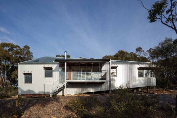 A compact single-storey house set within a wooded natural landscape. The exterior is clad in pale vertical corrugated metal, with a low-pitched corrugated metal roof, dark-framed windows, and a central elevated glazed balcony or living area accessed by an exterior stair. The building sits lightly above a rocky, bush-covered site and has a simple, utilitarian contemporary form.