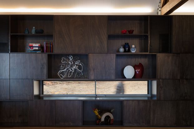 A dark timber built-in wall unit spans the image, combining closed storage cabinetry with open display niches, books, ceramics and artwork. A long horizontal glazed opening runs through the centre, while concealed linear lighting washes the upper wall. A substantial exposed timber beam is visible at the upper right, and warm natural light enters from outside.
