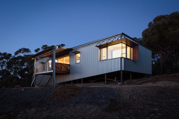 A modern elevated house photographed at dusk, set among trees on a sloping site. The compact rectangular building has corrugated metal cladding, large illuminated windows, a rooftop or upper-level railing, an external stair, and a timber-decked balcony. The structure is raised on slender posts above the rocky ground, creating a lightweight contemporary form.