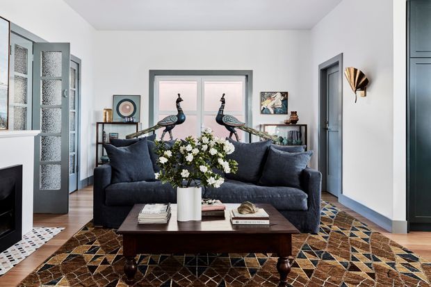 A refined living room with a deep navy-blue sofa, dark wood coffee table, patterned geometric rug, and a floral arrangement. White walls and a pale ceiling create a bright backdrop, while dark-framed glazed doors, a central window, built-in shelving, decorative peacock sculptures, artwork, and sculptural wall lighting add character. A fireplace is partially visible at the left edge.