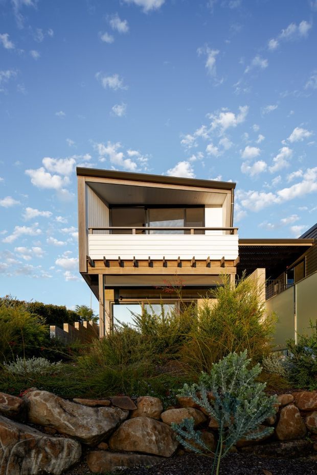 Exterior view of a contemporary multi-level home with a projecting upper-floor balcony, white horizontal balustrade, timber-lined soffits and framing, dark window and door frames, and broad glazing. Native-style drought-tolerant planting and natural stone retaining walls occupy the foreground beneath a clear blue sky.