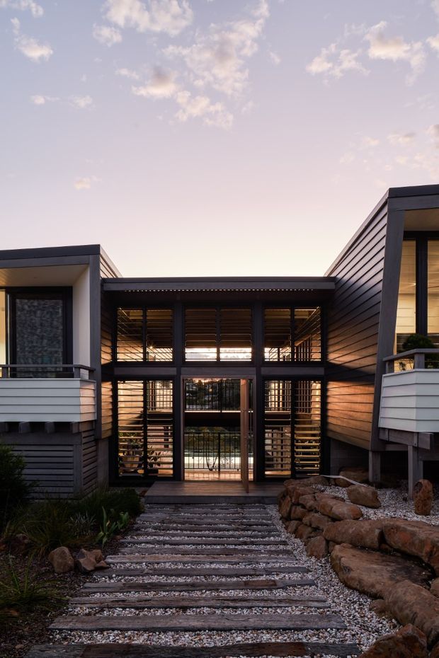 Exterior view of a contemporary split-level dwelling at dusk, with dark horizontal cladding, projecting upper volumes, and a glazed central entry bridge or breezeway. The entry is approached by a landscaped path of timber sleepers and gravel, with stone retaining edges and low planted areas. Strong geometric lines, extensive glazing, and warm interior lighting create a dramatic architectural composition.