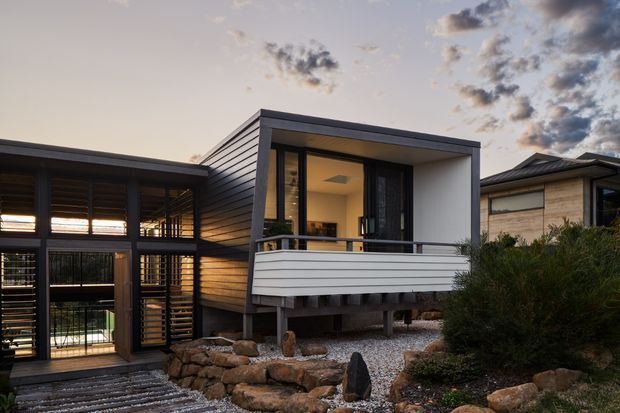 Exterior view of a compact contemporary residence composed of interconnected volumes. The main elevated pavilion has dark horizontal cladding, a projecting flat roof, expansive glazing, and a white horizontal-slatted balcony enclosure. A darker glazed linking volume with external sun-louvres sits to the left, while a neighbouring modern building is visible to the right. The home is set among rocks, gravel, and low landscaping, photographed in warm evening light.