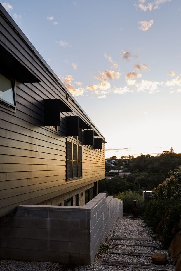 Exterior architectural photograph of a contemporary multi-level dwelling on a sloped suburban site at sunset. The facade is clad in dark horizontal weatherboards with projecting box-like window awnings and dark-framed glazing. A concrete block retaining wall and gravel stair path descend beside the building, with residential landscape visible beyond.