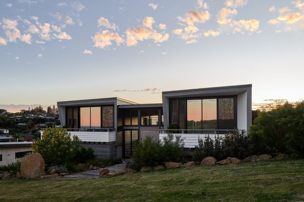 Exterior view of a contemporary single-storey home at sunset, composed of two offset rectangular wings with dark framed floor-to-ceiling glazing, textured dark masonry feature walls, light horizontal cladding, and a sheltered glazed central connection. The house sits on a landscaped, gently sloping site with rocks and planting, overlooking a suburban area.