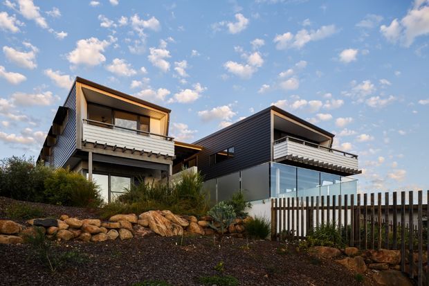 Exterior view of two adjacent contemporary multi-level homes on a landscaped, rocky site. The buildings feature dark horizontal cladding, contrasting light rendered or metal wall panels, projecting upper balconies with slatted privacy screens, large glazing, and asymmetrical pitched or skillion-style roof forms. A timber picket fence and native-style planting occupy the foreground beneath a blue, partly cloudy sky.