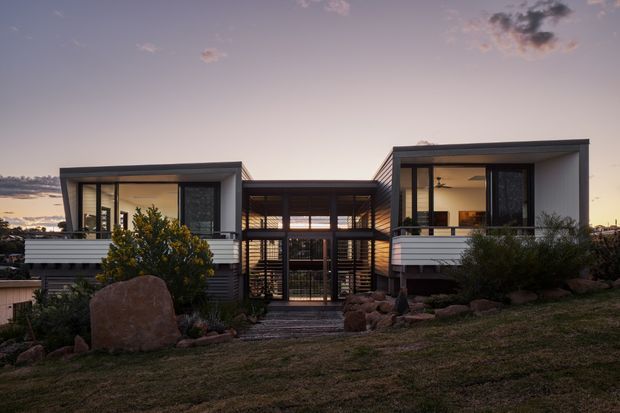 Exterior architectural photograph of a contemporary, low-rise residence at dusk. The house has two projecting box-like wings connected by a recessed central glazed entry, extensive dark-framed glazing, horizontal cladding, and a sheltered slatted central facade. It sits in a landscaped, rocky suburban setting with warm interior lighting visible through the windows.