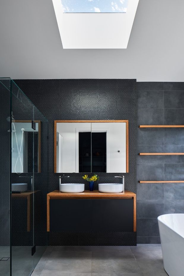 Contemporary bathroom with a double vanity, wall-mounted dark cabinetry, timber-edged mirror, black small-format mosaic tile feature wall, large dark grey wall tiles, grey floor tiles, a glass shower enclosure, freestanding bathtub, floating timber shelves, and a skylight providing natural light.