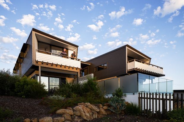 Exterior view of a contemporary multi-level residential building on a landscaped, sloping site. The composition shows two offset box-like volumes with dark horizontal cladding, light timber-toned balcony structures, projecting upper-level balconies, extensive glazing, and a glass balustrade. Native-style planting, rocks, and timber fencing occupy the foreground beneath a bright blue sky.