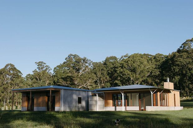 Single-storey contemporary house set in a wooded landscape, with a low-pitched metal roof, dark timber and grey wall cladding, extensive glazing, and a simple linear form. The building is viewed from the exterior across a grassy foreground.
