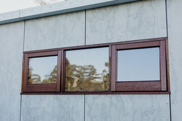 Close-up exterior view of a contemporary facade clad in large pale grey cementitious panels, with a horizontal arrangement of dark reddish-brown timber-framed windows. The central glazing reflects trees, while the side windows appear operable. A simple metal roof edge is visible at the top.