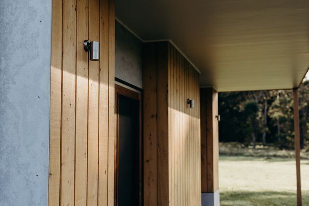 A close exterior view of a modern covered entry or verandah, featuring vertical natural timber wall cladding, dark recessed doorways, simple wall-mounted cylindrical lights, a smooth flat soffit, and a concrete or rendered wall edge. The covered walkway opens toward a grassy, wooded landscape.