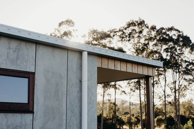 Close-up exterior view of a low-profile building with grey cement-fibre or concrete-panel cladding, a shallow metal roof, dark-framed window, white downpipe, and a covered timber-lined outdoor opening. Trees and a natural landscape are visible beyond the structure.