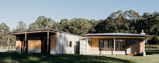 Single-storey contemporary rural residence composed of connected pavilion-like volumes, with dark corrugated metal roofing, timber-clad walls, light rendered or metal-panel sections, broad covered verandas, and large glazed openings. The house sits in a grassy, wooded setting with mature trees behind it.