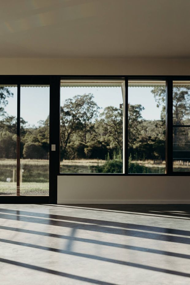 A largely empty, sunlit interior with expansive black-framed windows and glazed doors overlooking a wooded rural landscape. Strong linear shadows fall across a pale concrete or stone floor, while the walls and ceiling are plain and lightly coloured. The space has a clean, minimalist architectural character with generous glazing and a low, simple ceiling.