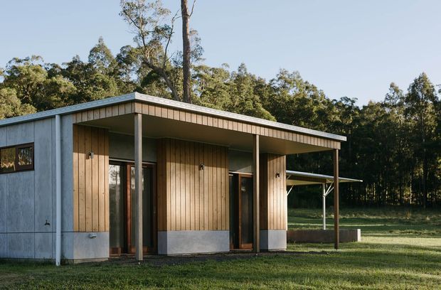 Single-storey modern cabin-like building set in a wooded rural landscape. The compact structure has a low-pitched flat roof with broad overhanging eaves, vertical timber wall cladding, pale concrete or masonry base walls, dark-framed glazing and slender timber posts. A covered carport or sheltered outdoor area extends to the right. The facade is photographed from outside across a grassy foreground, with dense trees behind.