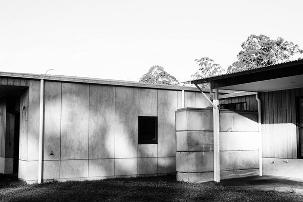Black-and-white photograph of a low, single-storey modern building with a flat or very shallow-pitched roof, vertical metal or fibre-cement wall panels, dark timber cladding, and a covered carport-like entrance. Mature trees are visible behind the structure.