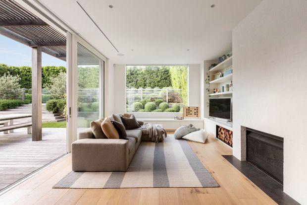 Bright contemporary living room with a low-profile sectional sofa, neutral striped area rug, pale timber flooring, built-in open shelving, and a minimalist dark fireplace set into a white wall. Large sliding glass doors open to a timber deck and landscaped garden, with additional garden views through the rear window.