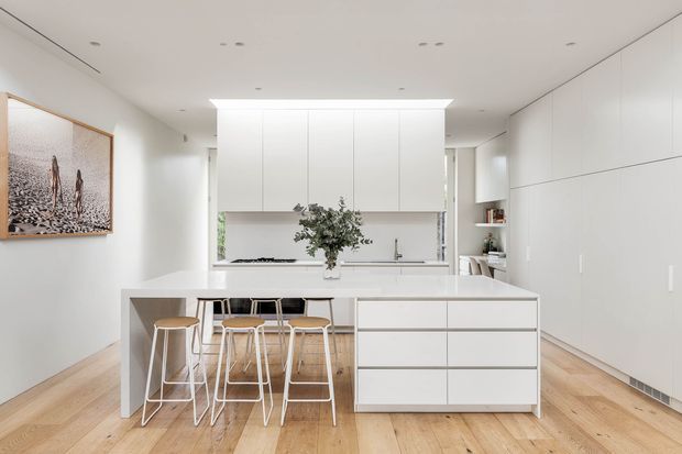 Bright, minimalist open-plan kitchen with a large white island incorporating seating and drawer storage. Full-height white cabinetry lines the right wall, while a central white kitchen run sits beneath a skylight-like ceiling opening. Pale timber flooring, recessed lighting, light wood stools, and a framed artwork create a clean, airy contemporary interior.