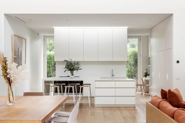 Bright open-plan kitchen and living area with a minimalist white kitchen, central island, timber flooring, light timber dining table, and warm rust-toned seating. Large windows bring in garden views, while concealed cabinetry and clean linear detailing create a refined contemporary interior.