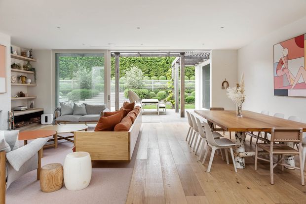 Bright open-plan living and dining area with pale timber flooring, a large timber dining table, upholstered dining chairs, layered sofas and armchairs, open shelving, and full-height glazing opening to a landscaped garden. The room has a clean contemporary feel with warm natural materials and colourful abstract wall art.