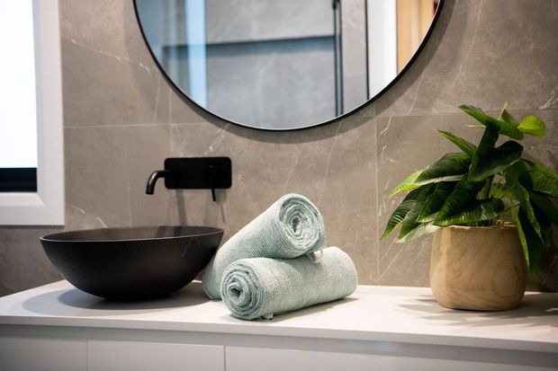 Contemporary bathroom vanity featuring a matte black vessel basin, wall-mounted black tapware, pale stone-look wall tiles, a large round mirror, white vanity cabinetry, rolled towels, and a potted plant.