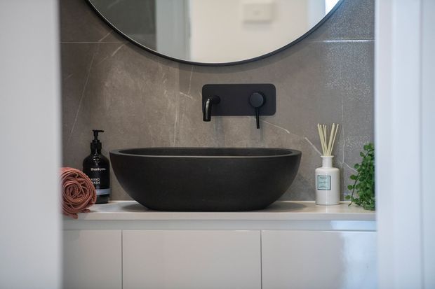 Close-up view of a refined bathroom vanity with a matte black vessel basin, black wall-mounted tapware, pale stone-look wall tiles, and white integrated cabinetry. A round mirror, soap dispenser, diffuser, and small plant complete the calm contemporary styling.