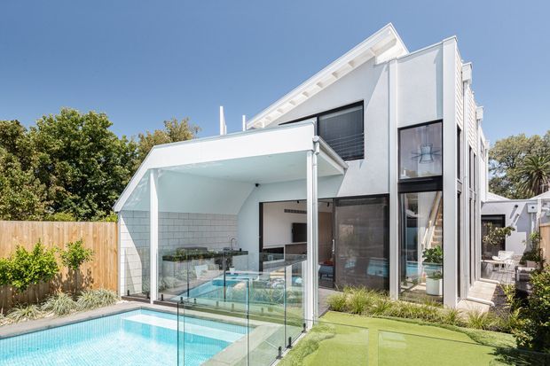 Exterior view of a contemporary multi-level residence with a white rendered facade, steep gabled roof, extensive floor-to-ceiling glazing, and a covered outdoor entertaining area. A swimming pool, glass balustrades, landscaped garden, and artificial turf courtyard occupy the foreground.