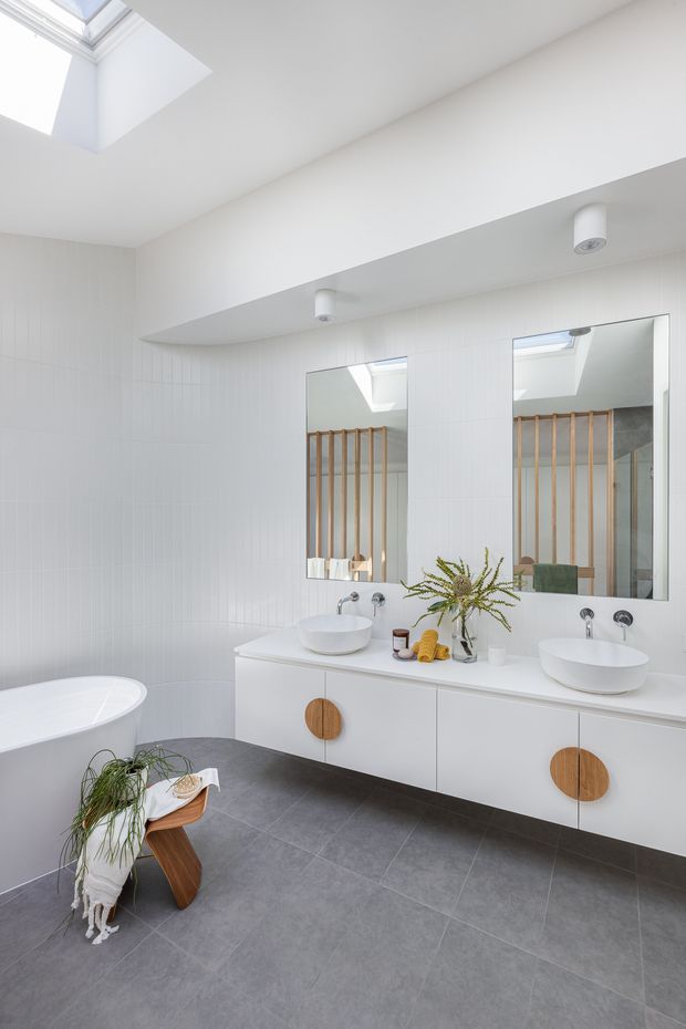 Bright contemporary bathroom with a freestanding white bathtub, double vanity, large frameless mirrors, white vertically textured wall tiles, and dark grey floor tiles. A skylight brings in abundant natural light, while timber privacy battens are reflected in the mirrors. The vanity features round timber handles and white vessel basins.