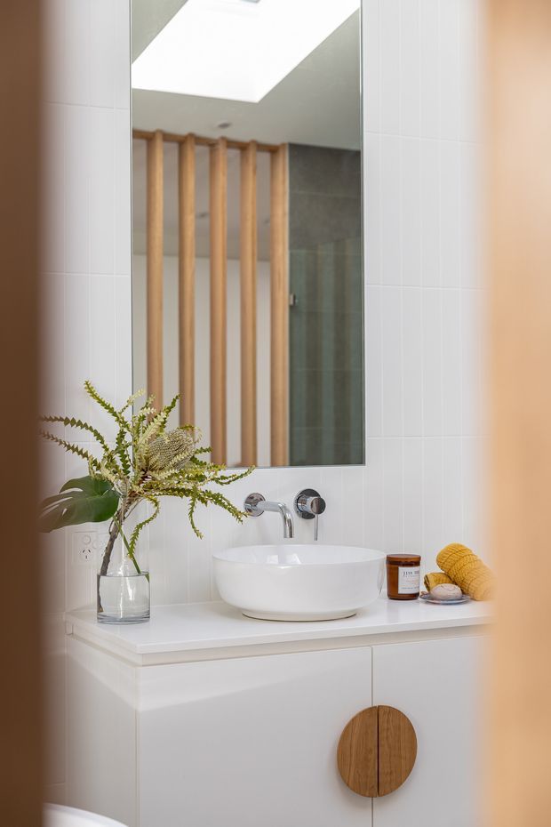 A bright, compact contemporary bathroom vanity with a white vessel basin on a white built-in cabinet. White vertically tiled walls surround a large frameless mirror, which reflects a skylight, grey tiled shower area, and a vertical timber screening feature. Brushed chrome wall-mounted tapware, light timber cabinetry detail, and a small vase of greenery add natural accents.
