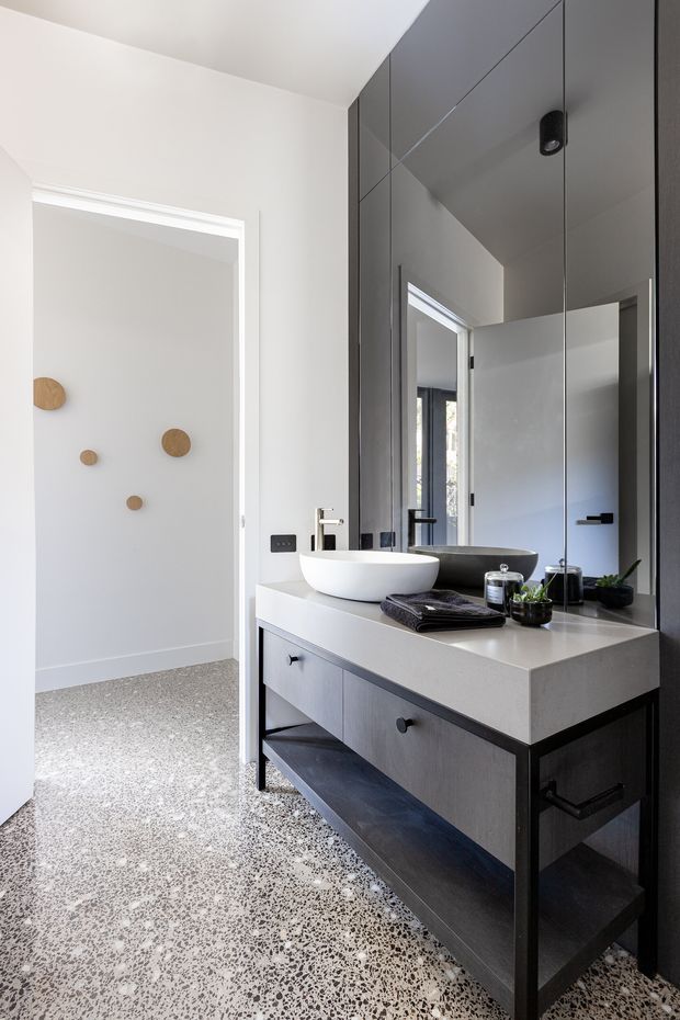Contemporary bathroom vanity with a white vessel basin, dark grey mirrored cabinetry, black tapware and a pale solid benchtop. A large frameless mirror spans the vanity wall, while terrazzo flooring extends into the adjacent doorway and white walls keep the space bright. A separate open doorway and subtle circular wall accents are visible to the left.