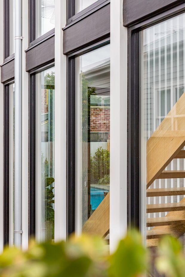 Close-up architectural view of a contemporary facade with tall, narrow glazed openings framed in dark charcoal and white vertical elements. A timber staircase is visible behind the glazing, with reflections and views toward a brick courtyard and blue swimming pool. The image emphasizes strong vertical lines, window framing, and the relationship between interior circulation and the outdoor courtyard.