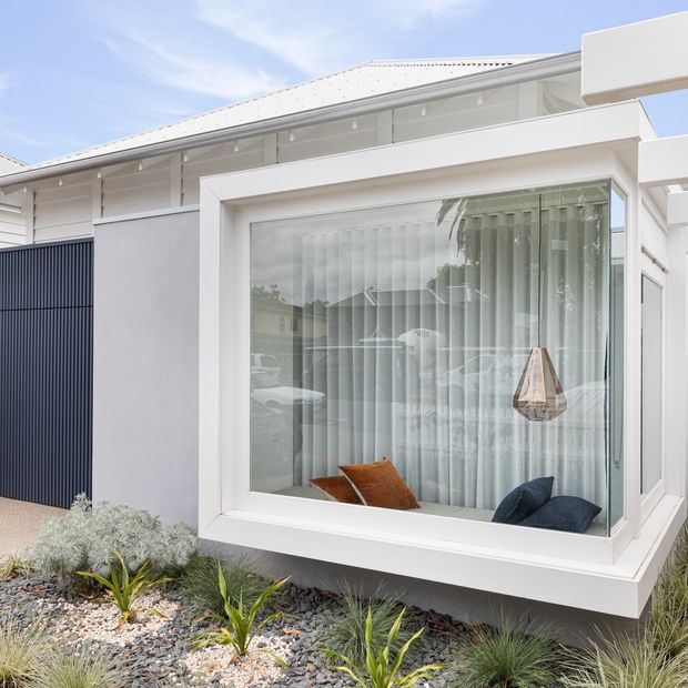 Bright exterior view of a contemporary white single-storey home, featuring a projecting box-bay window with floor-to-ceiling glazing and sheer curtains. A glimpse of a styled interior seating area is visible through the window, while the facade includes white cladding, dark vertical screening, and a light metal roof. The foreground has a low-maintenance landscaped garden with gravel and ornamental grasses.