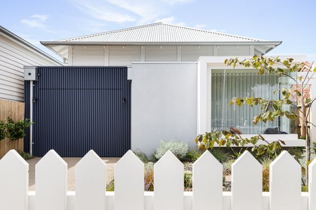 Bright street-facing view of a compact contemporary home with a pale rendered facade, low-pitched light metal hip roof, deep eaves, large window with sheer curtains, and a dark vertical-panel garage or entry door. A white picket fence and simple landscaped planting sit in the foreground.