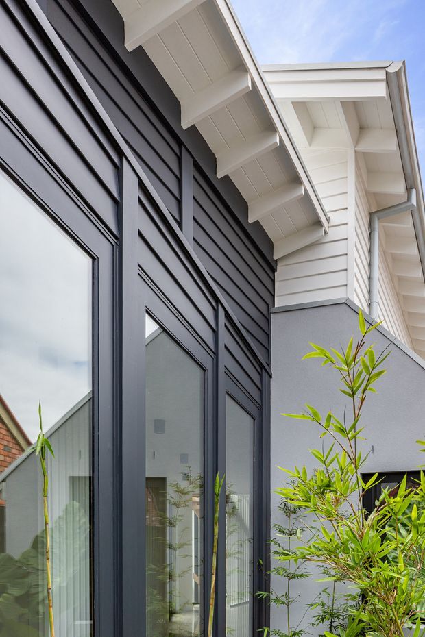 Close-up exterior architectural view of a contemporary home facade, showing dark vertical-framed glazing and dark horizontal cladding contrasted with white soffits, exposed eaves and light grey rendered wall surfaces. Bamboo-like planting is visible in the foreground, with reflected and partially visible interior space behind the glazing.