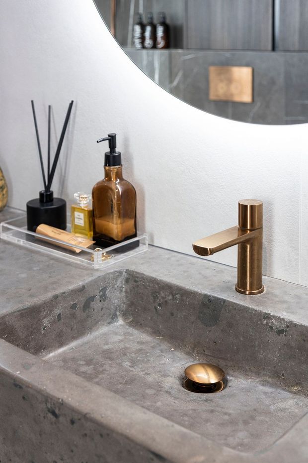 Close-up of a contemporary bathroom vanity featuring a deep integrated concrete or stone basin, a brushed bronze cylindrical tap, and a matching bronze drain cover. Amber soap bottles, a black reed diffuser, and toiletries sit on a clear tray against a textured white wall. A large rounded mirror reflects dark bathroom cabinetry and decor.