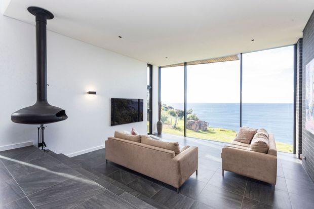 A bright, contemporary living room with floor-to-ceiling glazing overlooking the ocean. The room has a minimalist white ceiling and walls, dark large-format tiled flooring, a black freestanding suspended fireplace, two light beige sofas, a wall-mounted television, recessed downlights, and a dark exposed-brick feature wall. The expansive glazing provides a strong connection to the coastal landscape.