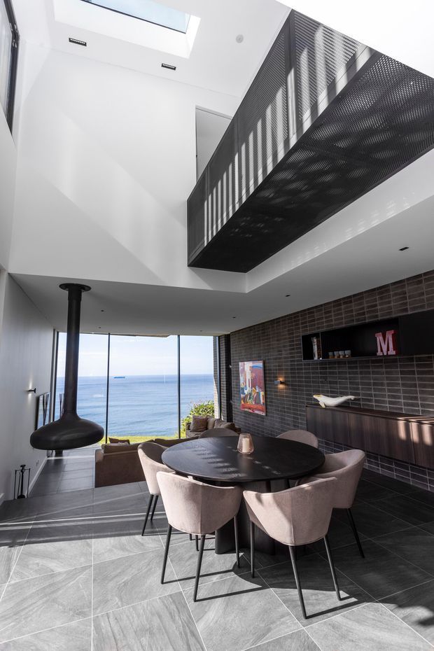 A dramatic double-height dining area overlooking the ocean through floor-to-ceiling glazing. The space features a round dark dining table with upholstered chairs, dark tiled flooring, a dark tiled feature wall with integrated cabinetry and shelving, a sculptural black suspended fireplace, and a perforated black mezzanine balustrade above. A skylight brings additional natural light into the otherwise contemporary interior.