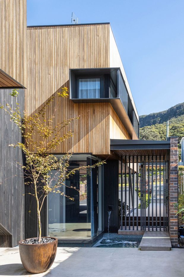 A contemporary multi-storey residential facade clad in vertical timber battens, with dark-framed glazing, a projecting upper-level window bay, a covered entry or carport-like structure, and a black vertical-bar gate. A shallow water feature sits beside the paved entrance, with a potted tree in the foreground. The building is photographed from an outdoor approach against a clear sky and wooded hillside.