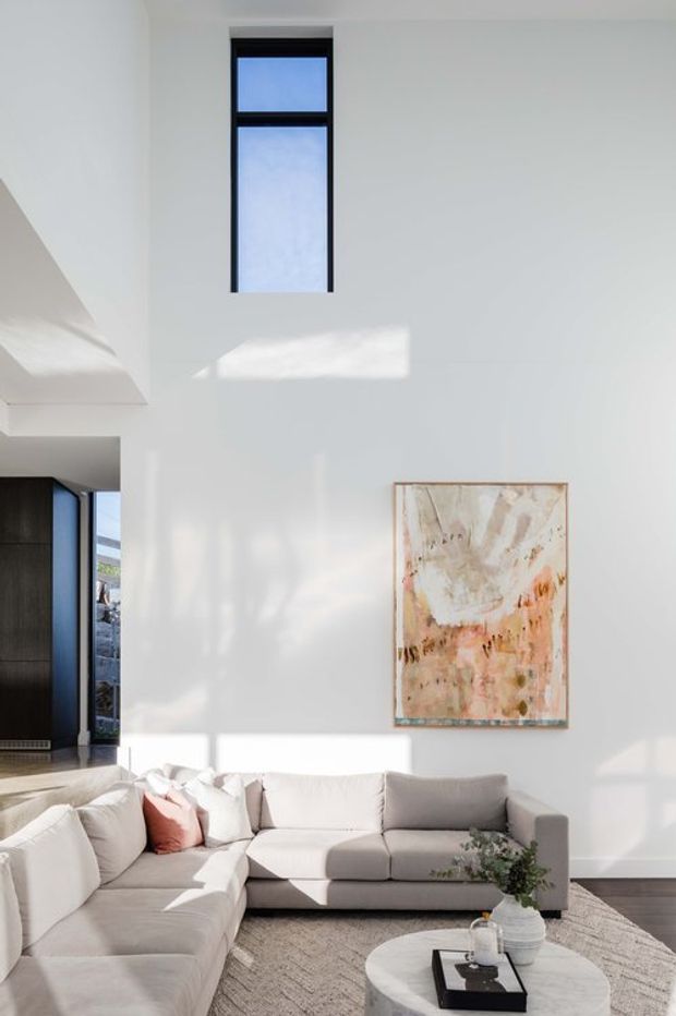 Bright double-height living room with white walls, a tall narrow black-framed window, abundant natural light, and a large light-grey sectional sofa arranged around a round coffee table. An abstract neutral-toned artwork hangs on the wall, with dark timber or stained joinery and glazing visible at the left. The space has a clean, refined contemporary feel.