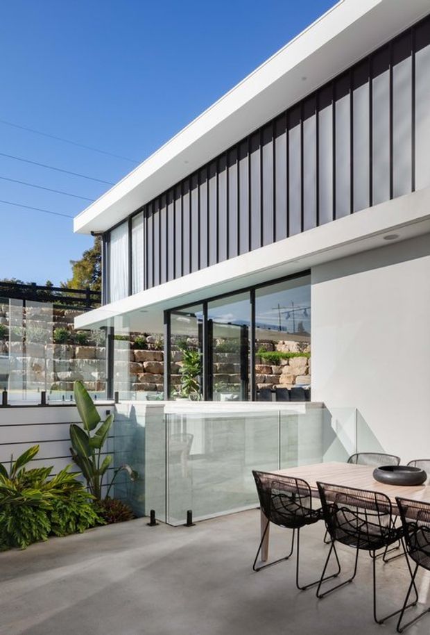 A bright contemporary two-storey home exterior viewed from an elevated outdoor terrace. The white rendered facade has a flat roof, extensive floor-to-ceiling glazing with dark slender frames, and a glass balustrade. A concrete terrace with a wire-frame dining table and chairs overlooks a landscaped lower courtyard and stone retaining walls, with lush planting and a clear blue sky.