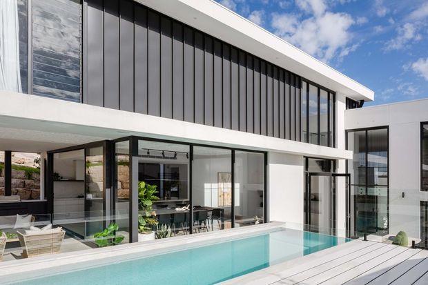 A bright contemporary multi-storey residence surrounding a turquoise swimming pool and pale timber deck. The facade combines crisp white rendered walls with extensive black-framed glazing and tall dark vertical cladding panels. Outdoor dining and lounge areas sit beneath covered terraces, with interior living spaces visible through the glazing. The composition emphasizes clean horizontal rooflines, strong geometric volumes, and indoor-outdoor connection.