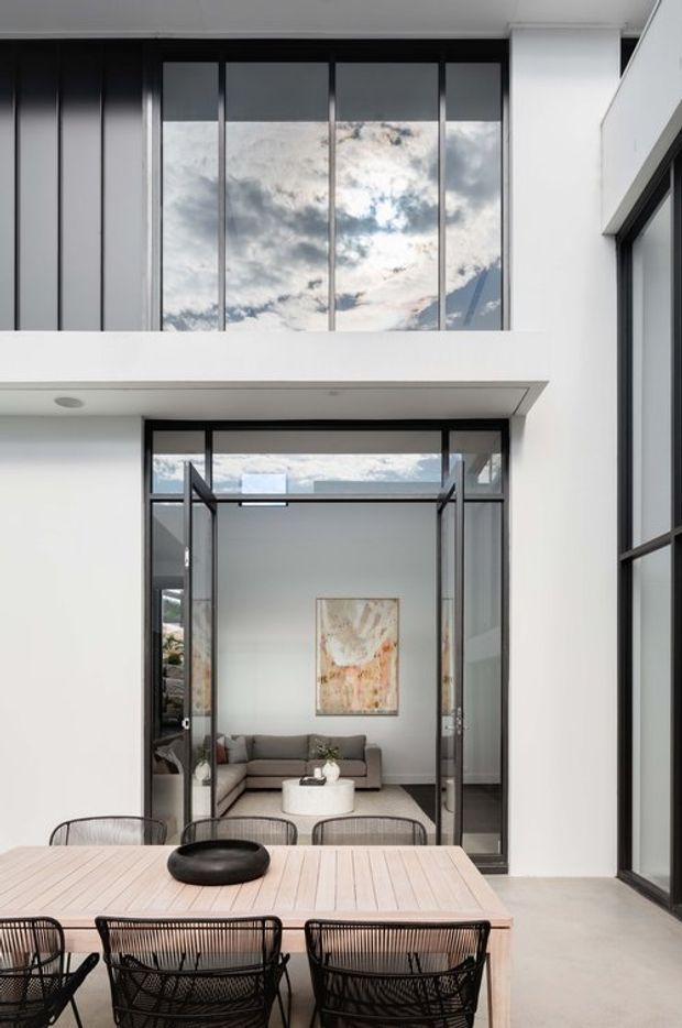 A contemporary multi-level courtyard-like space with a pale timber dining table and black wire chairs in the foreground. Large black-framed floor-to-ceiling glazing and open glass doors connect to a refined living room with a neutral sofa, round coffee table, and abstract artwork. White walls, concrete-toned flooring, strong vertical window divisions, and a high glazed void create a bright, airy indoor-outdoor composition.