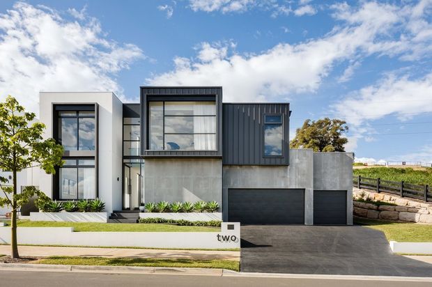 Contemporary multi-level residential facade with a strong geometric composition. The house combines smooth white rendered walls, exposed grey concrete, dark vertical metal cladding, large black-framed windows, and two dark garage doors. A broad paved driveway, landscaped front garden, retaining walls, and a crisp suburban street frontage are visible under a bright blue sky.