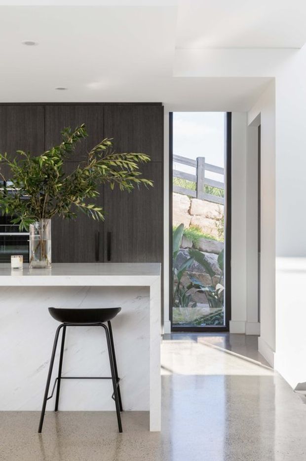 Bright contemporary kitchen interior with a white stone island and waterfall edge, dark timber-look full-height cabinetry, and a black bar stool. A narrow floor-to-ceiling glazed door or window opens toward a landscaped rock garden, while polished concrete or terrazzo-like flooring and recessed ceiling lights create a clean, minimalist feel.